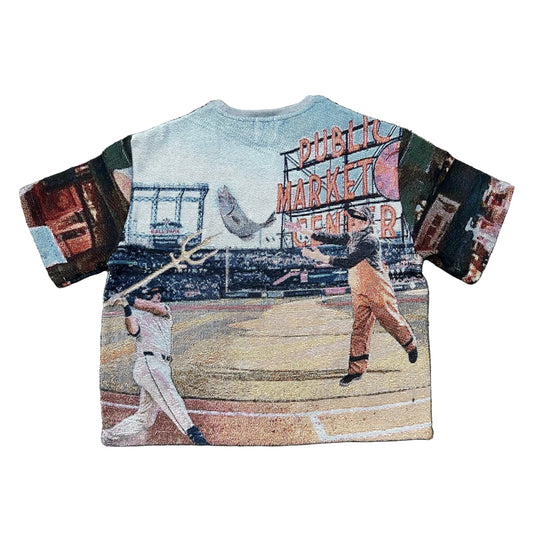 A short-sleeve woven tapestry T-shirt laid flat. The shirt features a baseball-themed collage print with a batter with trident, person throwing fish, stadium signage reading 'Public Market Center,' and a light blue sky area at the top.