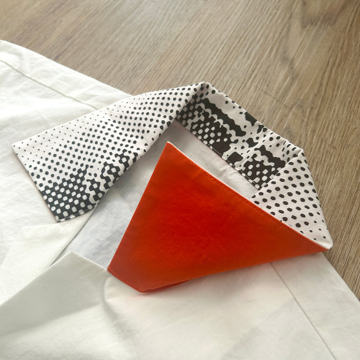 A folded white shirt with a patterned collar and a bright orange triangular fabric accent, placed on a light wood floor.