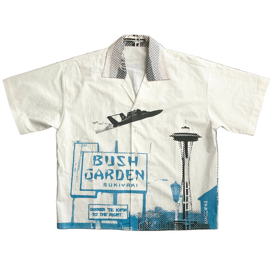 An off-white short-sleeve camp collar shirt featuring a blue and black graphic print of a hydroplane, the Seattle Space Needle, and a 'BUSH GARDEN' sign, shown flat against a white background.