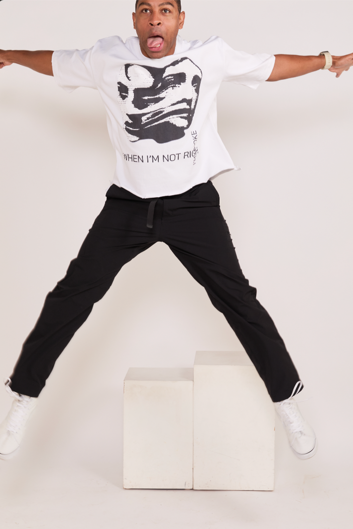 Person wearing a white t-shirt with a graphic design and text, black pants, and white shoes on a plain background.